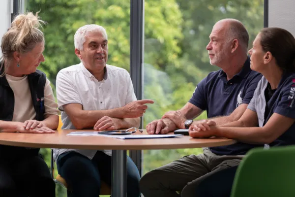 Drie zorgprofessionals en een patiënt zitten samen aan een ronde tafel bij het raam. De patient is een man van middelbare leeftijd met grijs haar. Hij draagt een spijkerbroek en wit overhemd. De professionals werken bij SVRZ, dit is te zien aan het logo op de kleding.