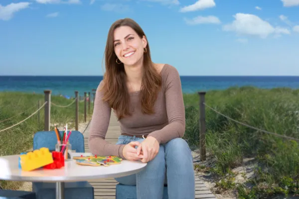 Foto van Liza Slager, Projectleider Kansrijke Start. De foto is samengevoegd uit twee bestanden. Liza zit aan een tafel met kinderspeelgoed. Deze setting is geplaatst tegen de achtergrond van een Zeeuwse omgeving. Op de achtergrond is een pad van houten planken te zien dat richting zee loopt. Vaag is een stukje strand en de zee te zien.