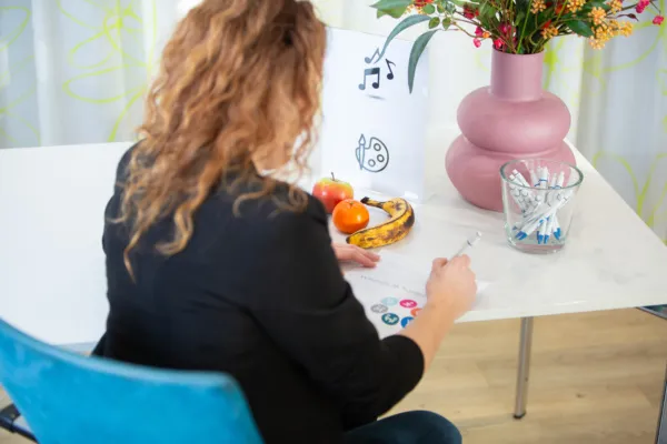 Afbeelding ter illustratie bij ketenaanpak Welzijn op recept. Op de afbeelding is een mevrouw met krullend haar op haar rug te zien. Ze zit aan een tafel en vult een formulier in. Op de tafel staat een glas met pennen, een vaas met bloemen en fruit.