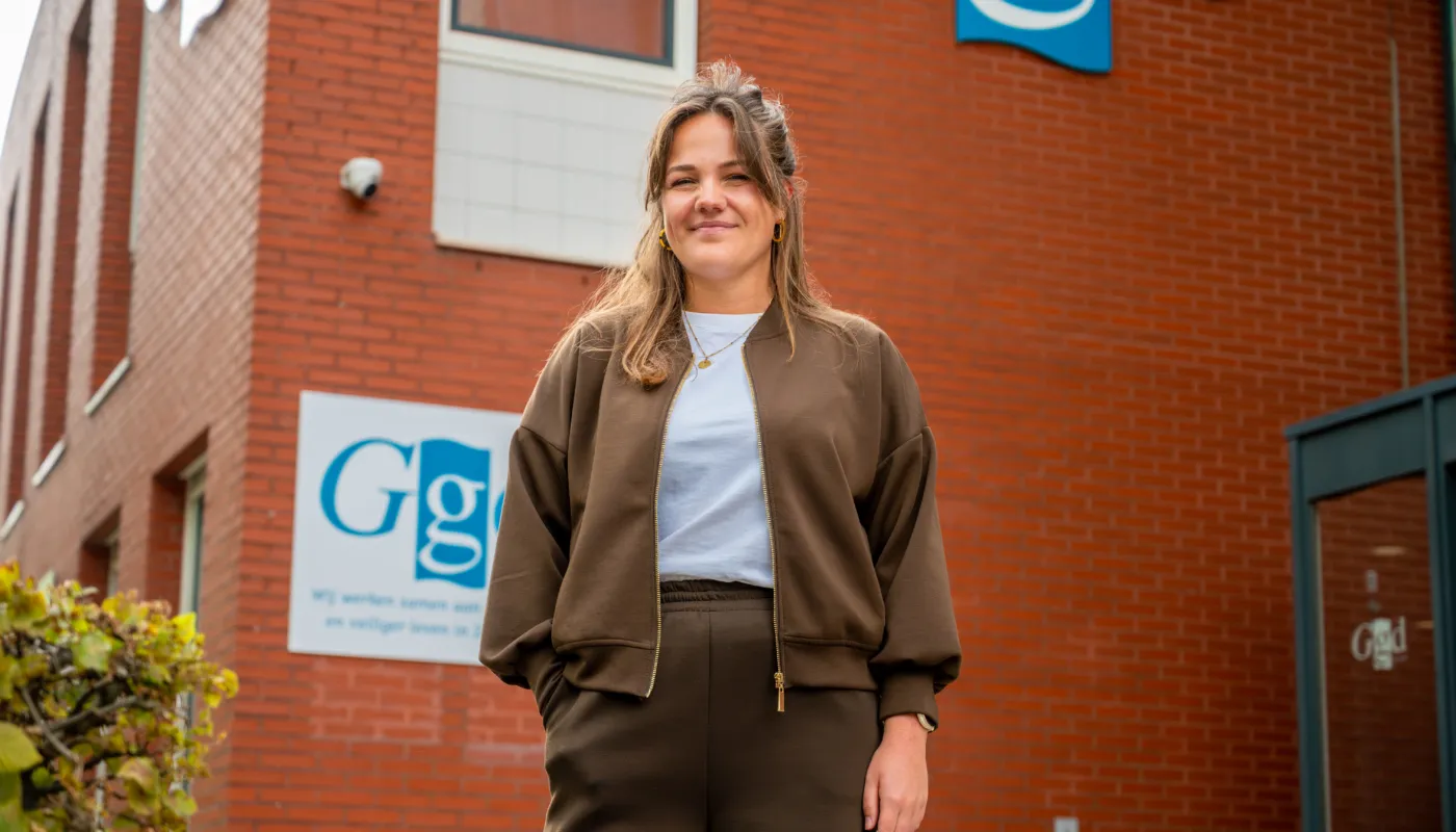Op deze foto is Iris Morauw te zien, staand voor het GGD-gebouw in Goes. Op de roodbruine bakstenen is tweemaal het GGD logo zichtbaar op de gevel. Ze draagt een bruine jas met rits en bruine broek, met daaronder een lichtgekleurd shirt.