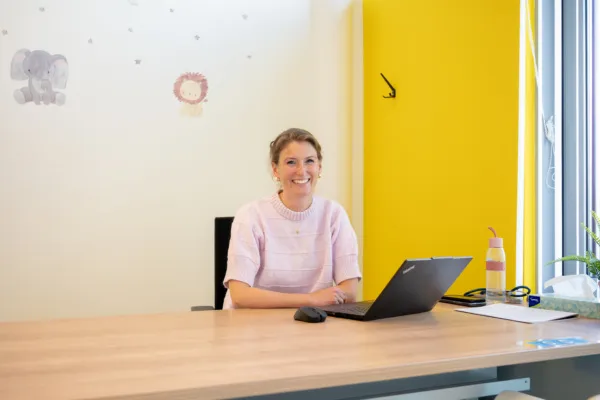 Op deze foto is Kristel Zevenbergen te zien. Ze zit aan een bureau in een kantooromgeving. Op het houten bureau staan een laptop, een muis en een waterfles. De achtergrond bestaat uit een witte muur met decoratieve stickers van een olifant, een leeuw en sterren. Aan de rechterkant van de foto is een felgele muur zichtbaar, en helemaal rechts is een raam te zien dat natuurlijk licht binnenlaat.