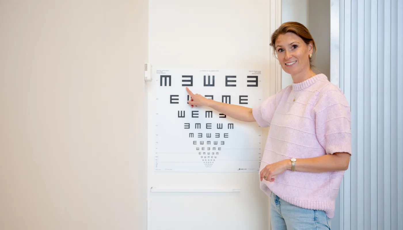 Op de foto is Kristel Zevenbergen te zien die naast een oogtestkaart staat. Ze wijst naar de bovenste rij van de kaart, waarop grote letters staan. De letters op de kaart worden kleiner naarmate je verder naar beneden kijkt. Ze draagt een lichtroze trui en een blauwe spijkerbroek. De ruimte heeft lichte muren en aan de rechterkant zijn verticale jaloezieën zichtbaar.