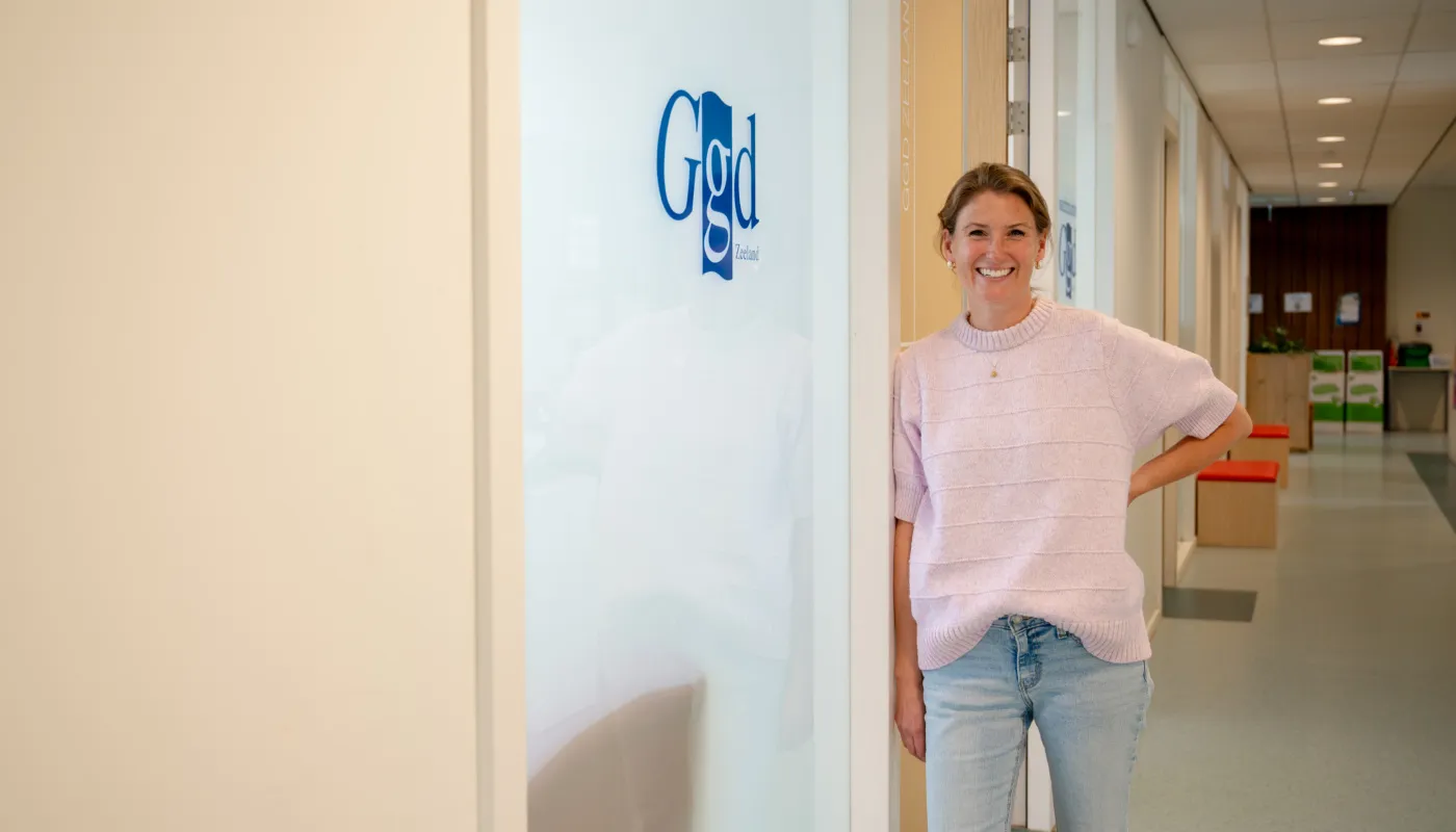 Op deze foto staat Kristel Zevenbergen in een gang naast een deur waarop het logo van GGD Zeeland te zien is. Ze leunt tegen het deurkozijn en draagt een lichtroze trui en blauwe spijkerbroek. In de achtergrond zijn houten bankjes met oranje kussens en een balie zichtbaar.