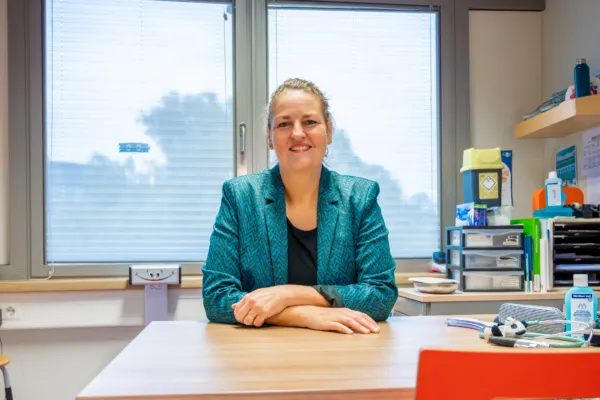 Op deze foto is Noor Adriaanse te zien, zittend aan een bureau in een consultatiebureau. Ze draagt een blauwgroene blazer over een zwart shirt en kijkt glimlachend in de richting van de camera. Op het bureau staan diverse items, waaronder handgel en kantoorartikelen. Achter haar zijn twee grote ramen waardoor natuurlijk licht binnenvalt. Rechts in beeld zijn planken te zien met boeken en kantoorbenodigdheden.