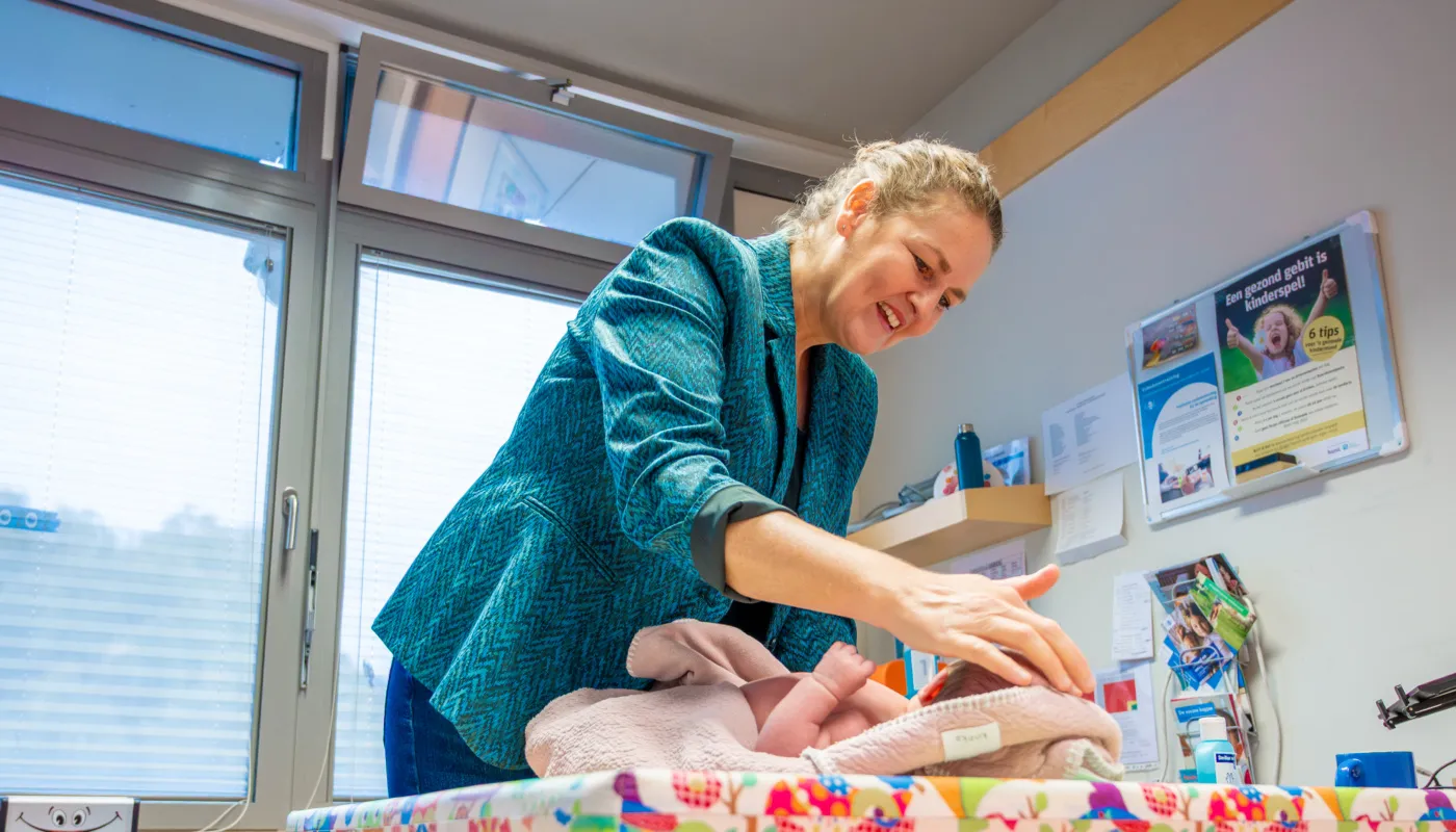 Op deze foto is Noor Adriaanse te zien terwijl zij een baby onderzoekt op het consultatiebureau. Ze draagt een blauwgroene jas en heeft haar haar vastgebonden. De baby ligt op een kleurige mat. Op de achtergrond zijn ramen met deels gesloten jaloezieën, en aan de muur hangen posters en flyers. Er is ook een wandplank met enkele spullen erop.