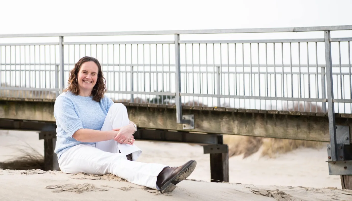 Foto van Margriet van Schelven, zittend op het strand. Ze zit ontspannen op het zand met gekruiste benen en leunt iets achterover. Ze dragen een lichtblauwe trui, witte broek en bruine schoenen. Op de achtergrond is een metalen railing zichtbaar die verhoogd boven het zand loopt. De omgeving oogt natuurlijk en kalm, met neutrale tinten die bijdragen aan de rustige sfeer van de scène.