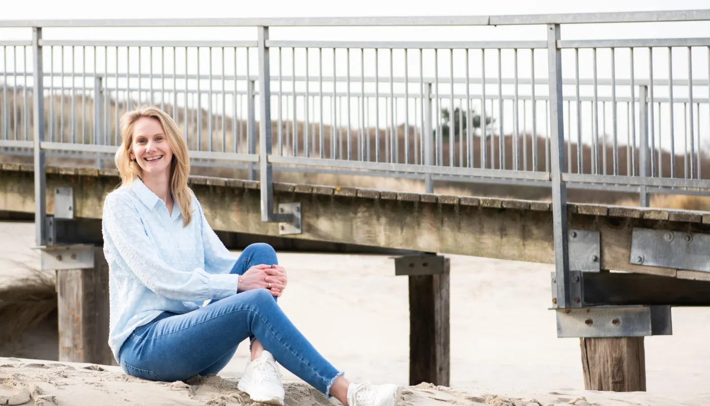 Op deze foto zit Miriam op een zanderige ondergrond in de buurt van een houten brug met metalen relingen. Ze draagt een lichtblauwe blouse met lange mouwen, een blauwe spijkerbroek en witte sneakers. Op de achtergrond is een brug zichtbaar, evenals wat vegetatie in de verte. De setting oogt natuurlijk en ontspannen. De foto is genomen in een Zeeuws duingebied.