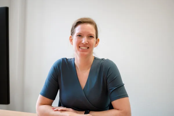 De foto toont Jenny Brouwer die aan een bureau met laptop zit in een eenvoudige, lichte ruimte. De houding is rechtop met beide armen ontspannen op het bureau. De kleding bestaat uit een donkerblauwe top met korte mouwen en een overslagdetail aan de voorkant. Aan de pols is een smartwatch zichtbaar. De achtergrond is minimalistisch, met een effen lichte muur.