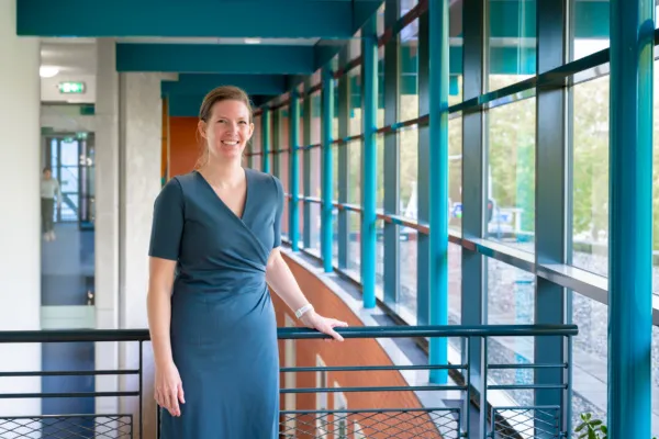 De foto toont Jenny Brouwer, staand in een moderne, lichte gang met grote ramen die veel daglicht binnenlaten. De ruimte heeft een strak en open ontwerp met blauwe balken en metalen leuningen langs een balustrade. De persoon draagt een elegante, donkerblauwe jurk met korte mouwen en een overslagdetail, en rust met één hand op de railing. Buiten is groen zichtbaar door de ramen.