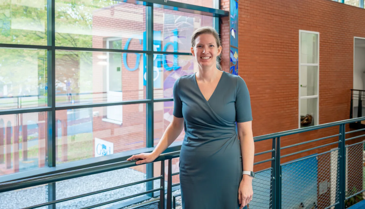 De foto toont Jenny Brouwer, staand in een moderne gang met grote ramen die veel daglicht binnenlaten. De achtergrond bestaat uit een rode bakstenen muur en een glazen wand met blauwe accenten en een kleurrijke print. Ze leunt licht tegen een metalen railing en draagt een elegante, donkerblauwe jurk met een overslagdetail en korte mouwen. Aan de pols is een smartwatch zichtbaar. De setting oogt ruim en licht, met een combinatie van industriële en moderne elementen.