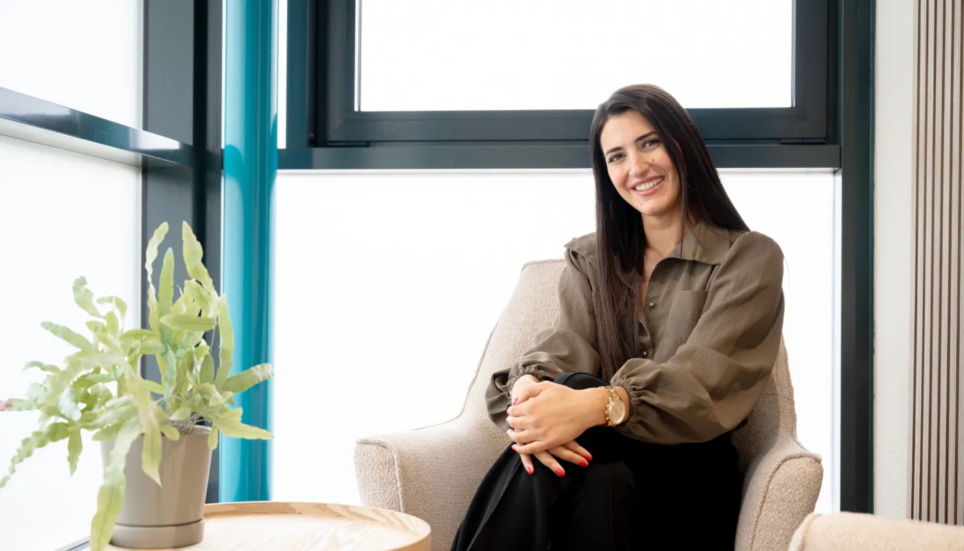 Op de foto zit Ozlem ontspannen in een lichtgrijze fauteuil met de benen gekruist en handen op de knie. Op de voorgrond staat een houten bijzettafel met een groene plant in een pot. Ze zit in een hoek met grote ramen, waardoor de ruimte heel licht is. Ze draagt een lange blouse in een neutrale kleur met pofmouwen en een zwarte broek.