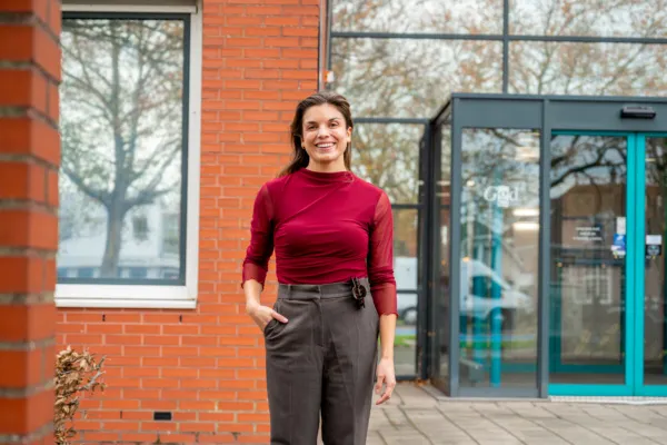 De foto toont Maureen Gonzales staand buiten voor de GGD hoofdlocatie. Het gebouw heeft een glazen ingang waarop “GGD” zichtbaar is. Ze draagt een donkerrode top met lange mouwen en een grijze, hoge taillebroek. Op de achtergrond zijn een rode bakstenen muur, een raam en een deur met blauwe kozijnen te zien, evenals enkele bomen buiten.